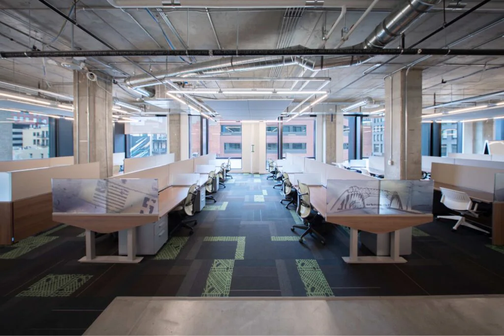 Rows of workstations with mesh task chairs and printed privacy panels beneath a concrete ceiling with floor-to-ceiling windows