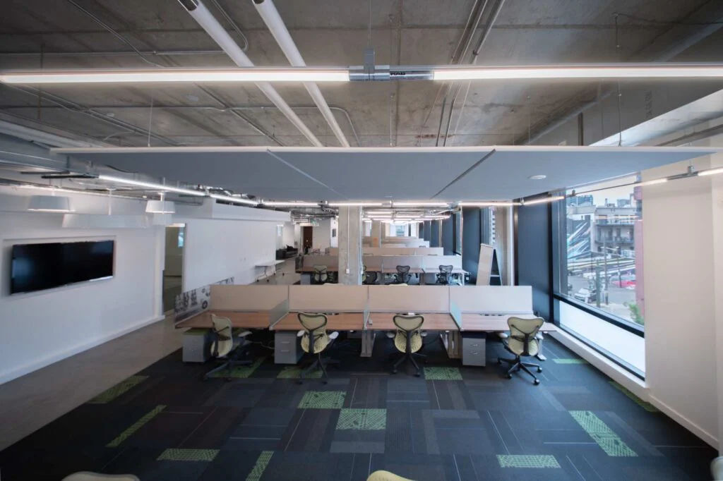 Open office bench seating with green task chairs, exposed concrete columns, and city views through large windows