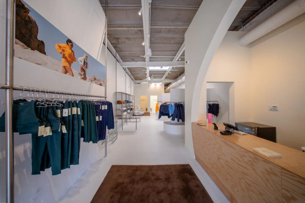 Store interior view down the aisle toward the glass entrance door past clothing racks and checkout counter