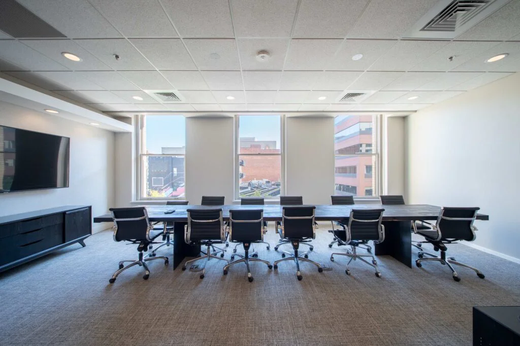 Large conference room with a dark wood table, leather chairs, a wall-mounted TV, and floor-to-ceiling city view windows