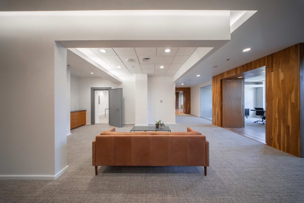 Lounge area with a tan leather sofa and glass coffee table positioned between hallways leading to private offices