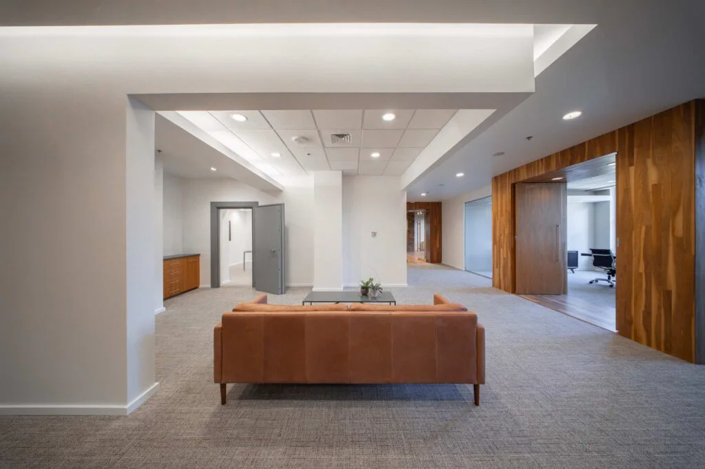 Lounge area with a tan leather sofa and glass coffee table positioned between hallways leading to private offices