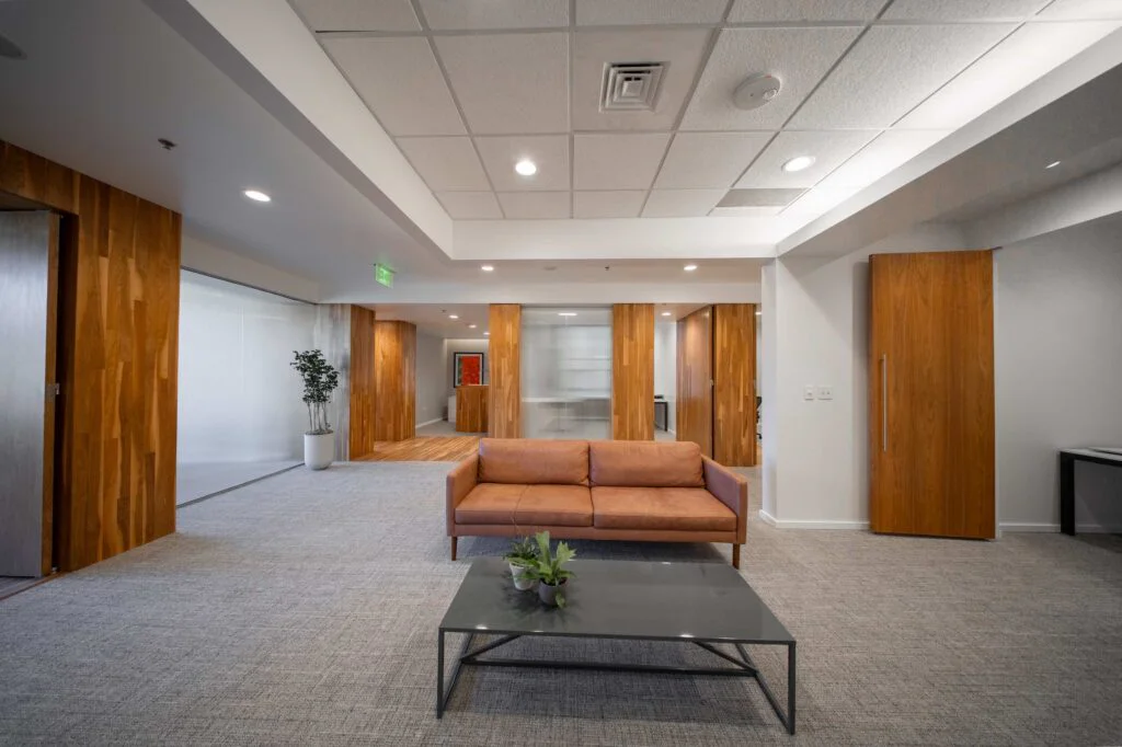 Reception lounge with a leather sofa, glass coffee table, potted plant, and wood-paneled doorways to private offices
