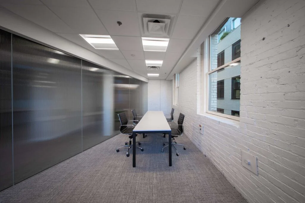 Office corridor with a glass-front meeting room, painted brick wall, and a window overlooking a neighboring building