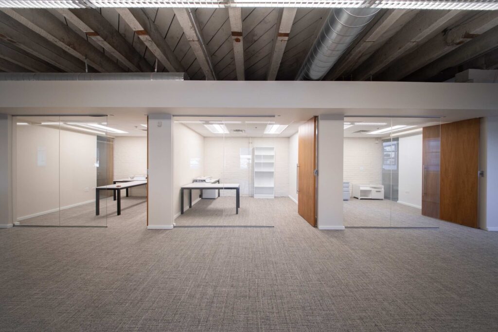 Central open area flanked by glass-walled offices with desks, separated by a wood-clad pillar and wood door