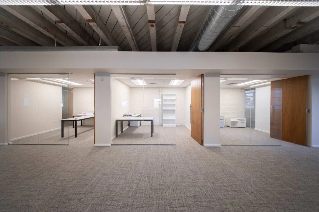 Central open area flanked by glass-walled offices with desks, separated by a wood-clad pillar and wood door