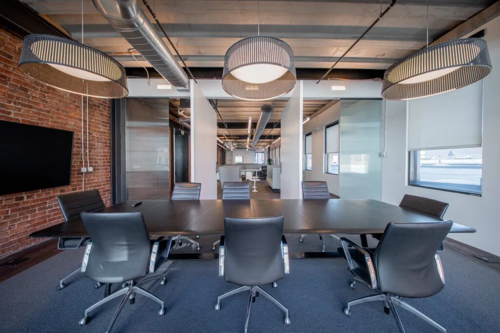 Conference room with pendant lights and a brick accent wall, opening onto a hallway with lounge seating and desks