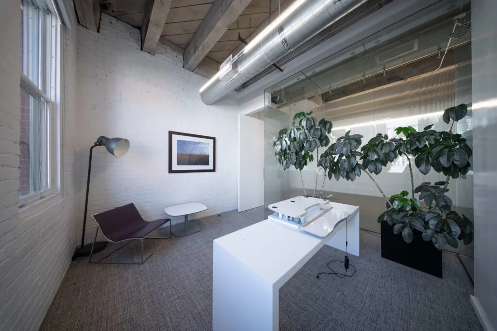 Office corner with a leather lounge chair, floor lamp, framed photo, and a white standing desk beside potted plants