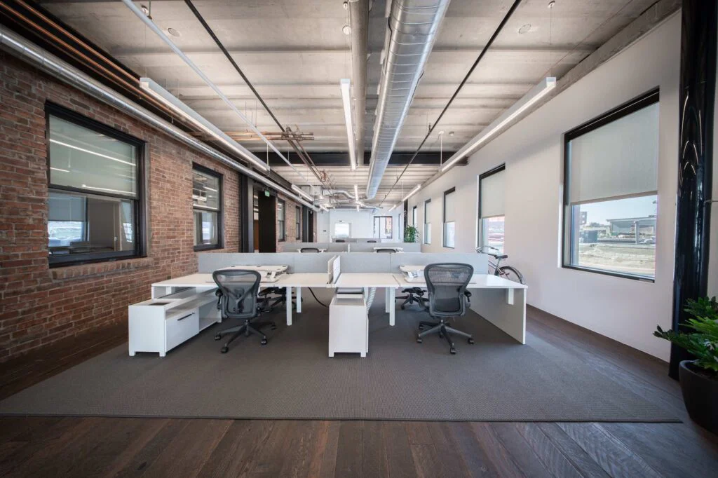 Open-plan desks with ergonomic chairs and privacy panels set against an exposed brick wall, with a bicycle parked nearby
