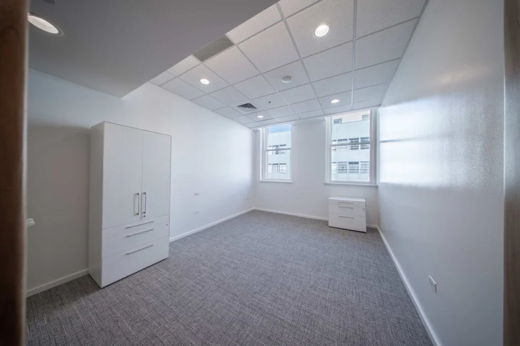 Unfurnished private office with a tall white storage cabinet, carpet tile flooring, and two windows overlooking downtown