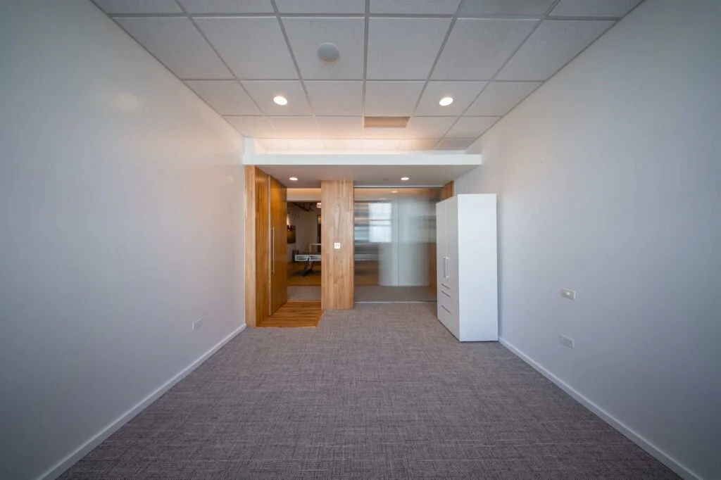 Carpeted hallway leading through an open wood door into an office with a white storage cabinet and glass partition