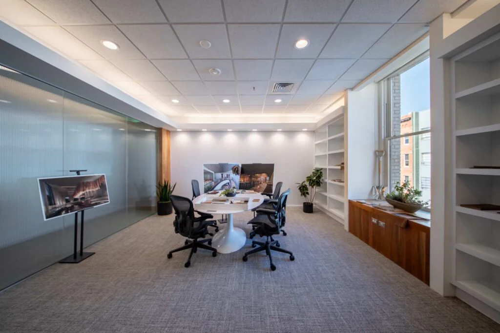 Design meeting room with a round table of fabric and material swatches, built-in bookshelves, and a display screen