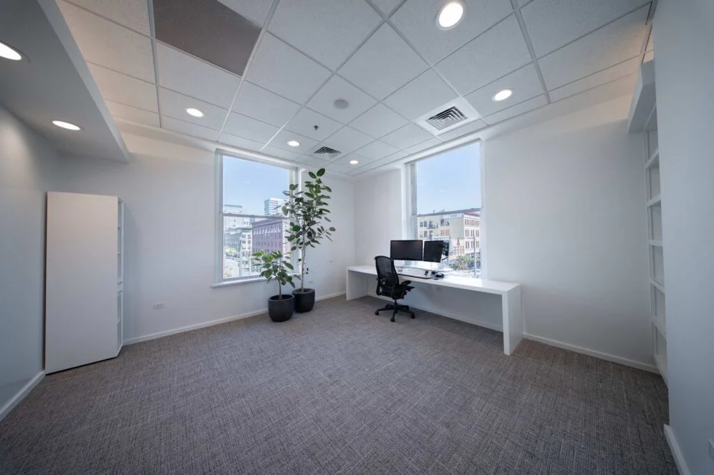 Private office with a white built-in desk, dual monitors, potted plants, and windows overlooking downtown buildings