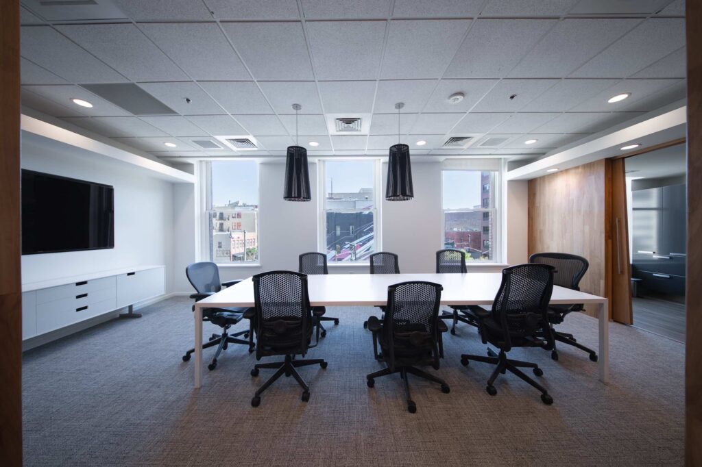 Bright conference room with black cone pendant lights, a wall-mounted TV, and windows overlooking the downtown skyline