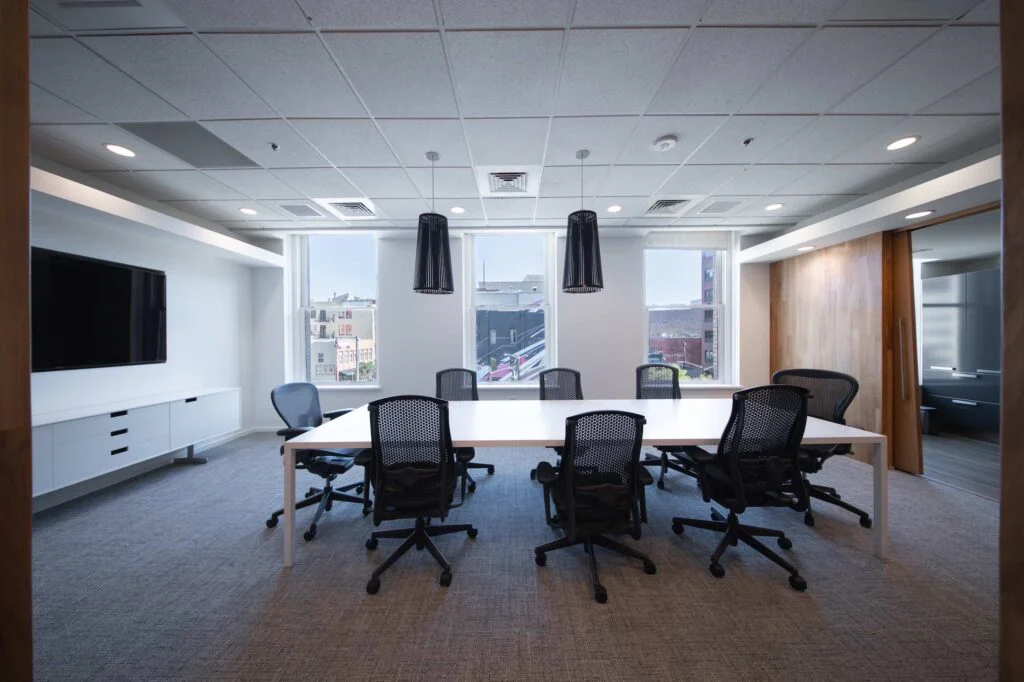 Bright conference room with black cone pendant lights, a wall-mounted TV, and windows overlooking the downtown skyline