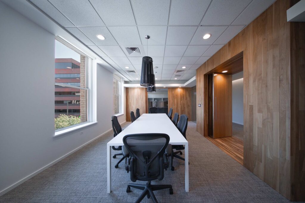 Long conference table with a black pendant light, opening through a wood-framed doorway to the office kitchen beyond