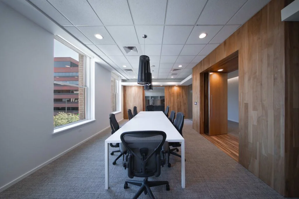 Long conference table with a black pendant light, opening through a wood-framed doorway to the office kitchen beyond