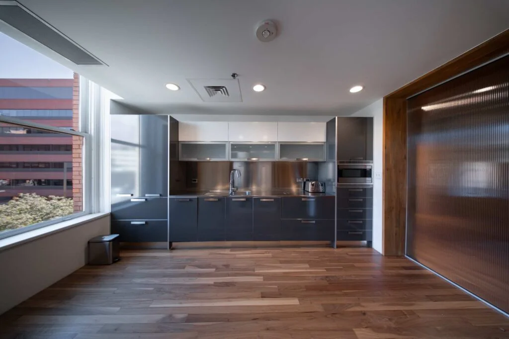 Office kitchen with dark gray cabinetry, stainless steel backsplash and countertop, and wood flooring
