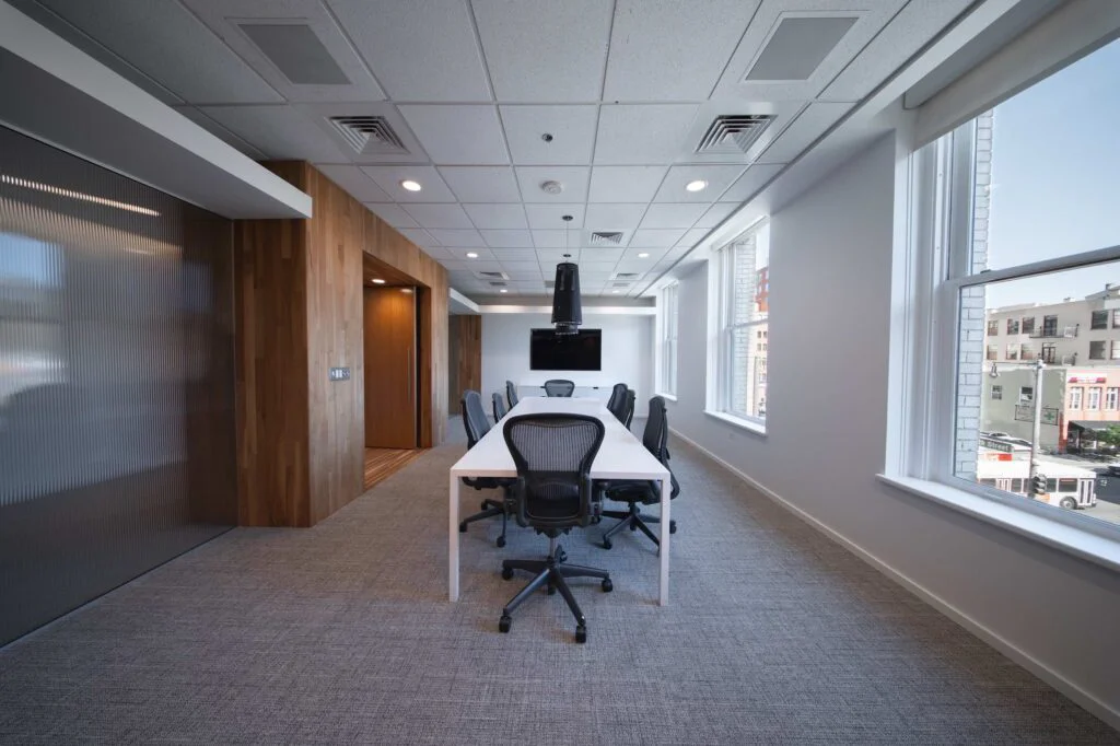 Long conference table and mesh chairs in a wood-paneled meeting room with floor-to-ceiling windows overlooking downtown street