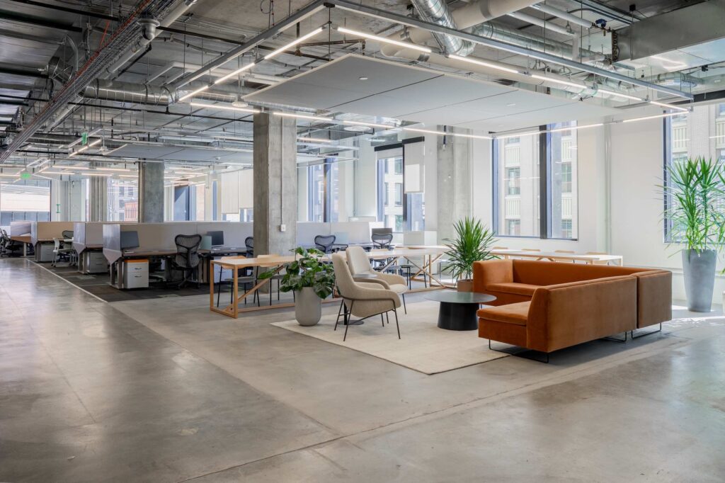 Office breakout lounge with an orange sectional sofa, standing desks, and floor-to-ceiling windows