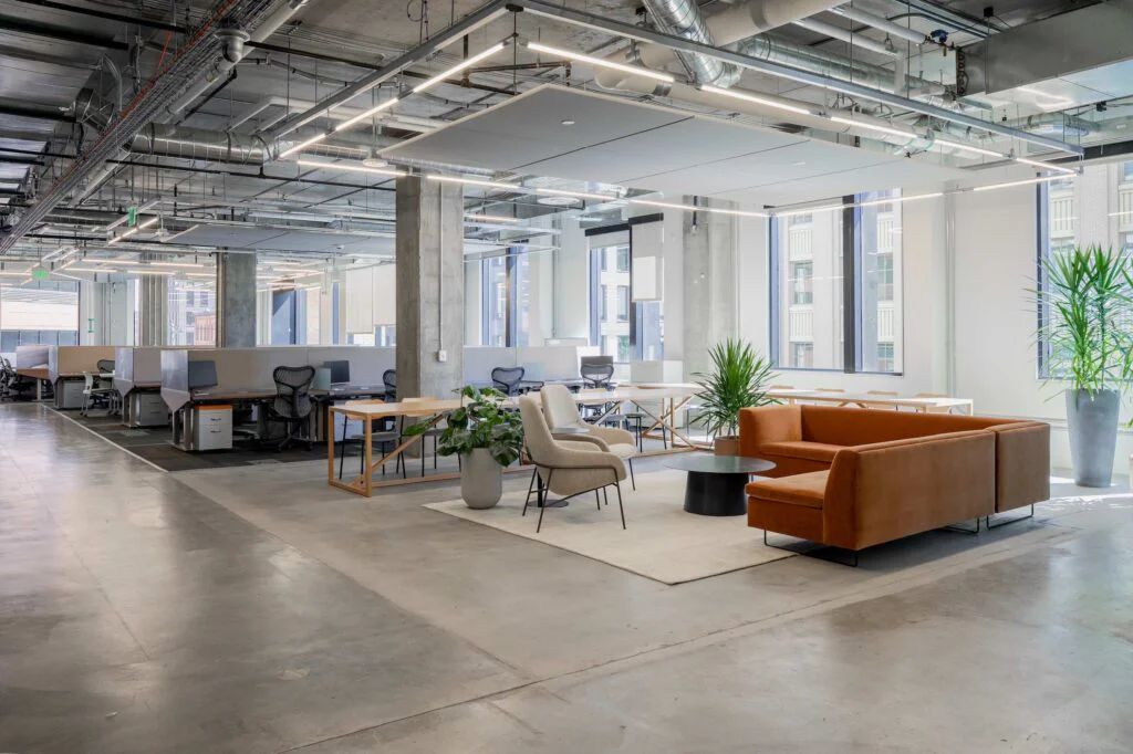 Office breakout lounge with an orange sectional sofa, standing desks, and floor-to-ceiling windows