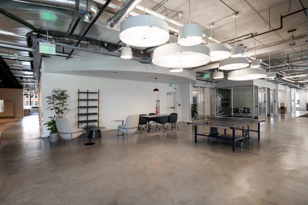 Office lounge with a ping pong table, lounge chairs, and a small meeting table near glass-walled offices