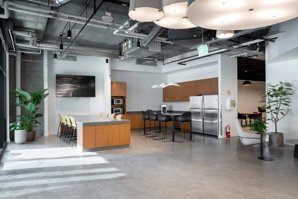 Break room kitchen with a wood island, black bar stools, wall-mounted TV, and stainless refrigerator