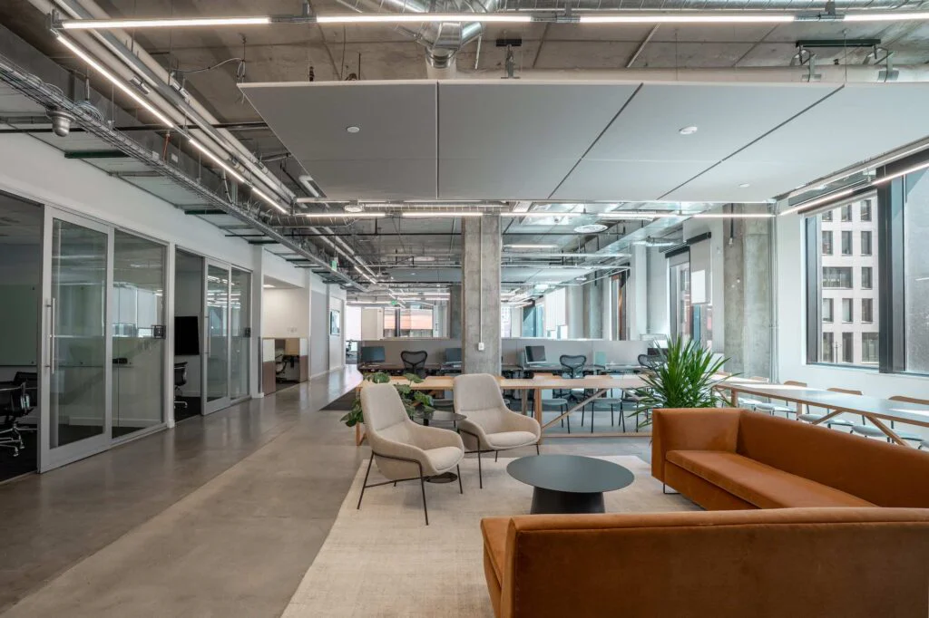 Hallway past glass-walled offices leading to a wood communal table and sectional sofa seating area