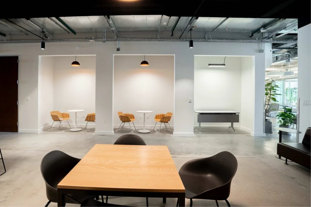 Wall alcoves with wood lounge chairs and round tables beside a small enclosed meeting room