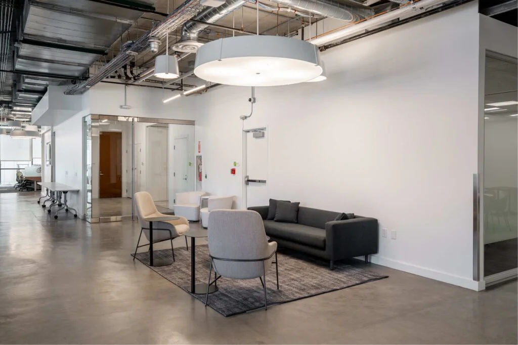 Gray sofa and accent chairs on a rug near glass-walled office entrances and a round side table