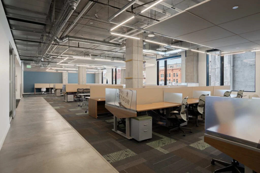 Rows of office desks and cubicles with a Detour sign, exposed concrete columns and city views