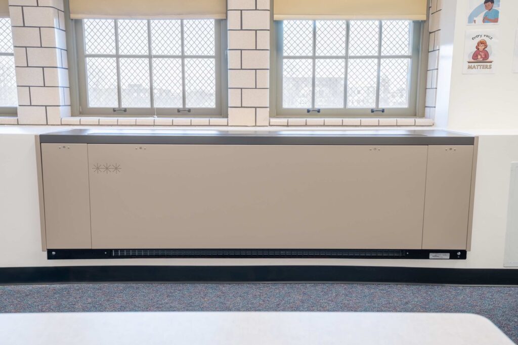 Beige radiator cover installed below classroom windows with a wall poster nearby