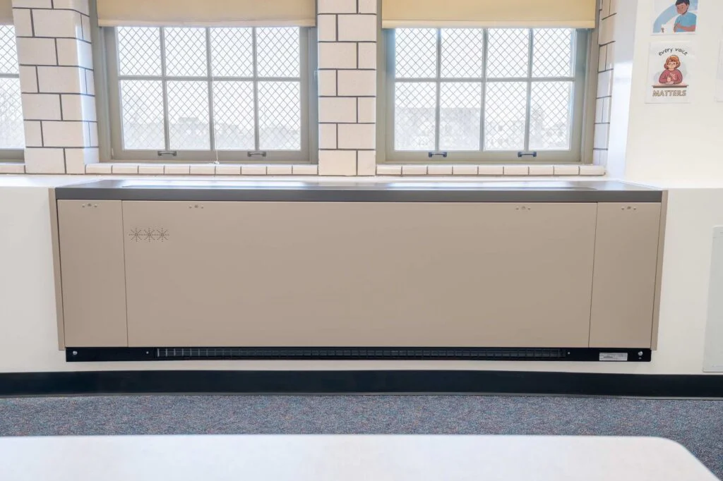 Beige radiator cover installed below classroom windows with a wall poster nearby