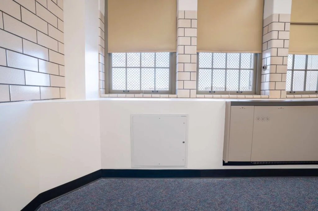 Wall access panel beside a radiator cover under classroom windows with security screens