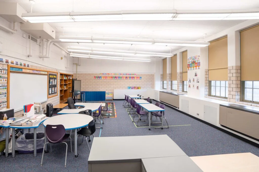 Wide view of a finished classroom with whiteboard, bookshelf, desks, and windows