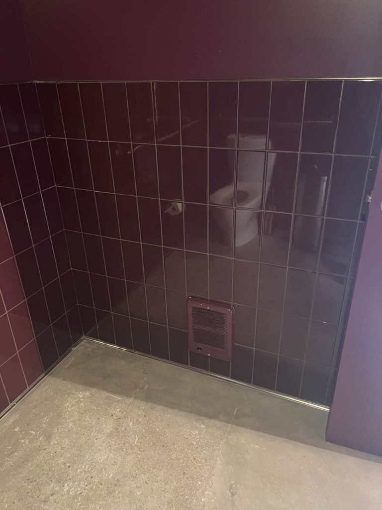Glossy maroon tiled restroom corner with an exhaust vent and toilet reflected in the mirror