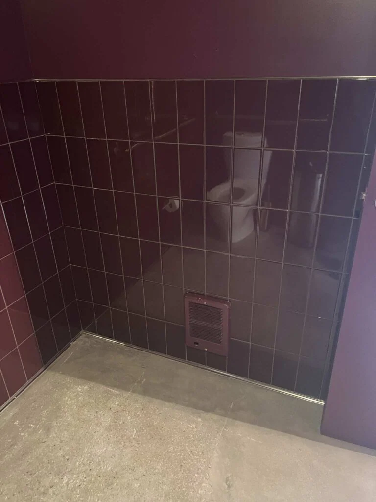 Glossy maroon tiled restroom corner with an exhaust vent and toilet reflected in the mirror