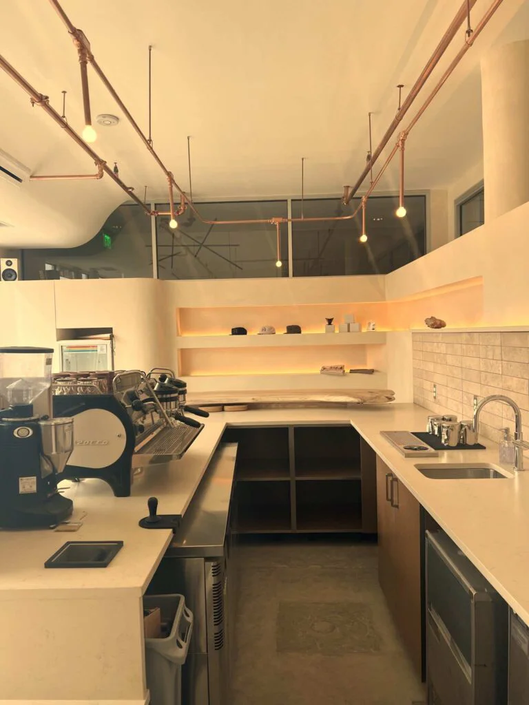 Wide view of a coffee shop bar with espresso equipment, backlit shelving, and copper pipe lighting overhead