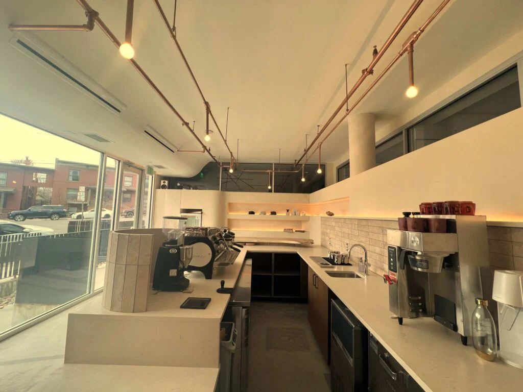 Exposed copper pipe lighting track running above a coffee counter with street-facing windows
