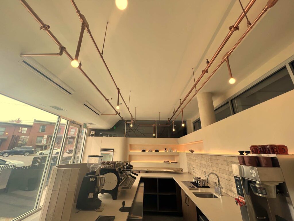 Copper pipe track lighting suspended above a coffee shop counter near large storefront windows