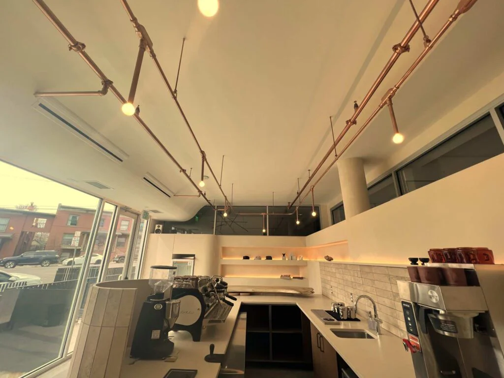 Copper pipe track lighting suspended above a coffee shop counter near large storefront windows