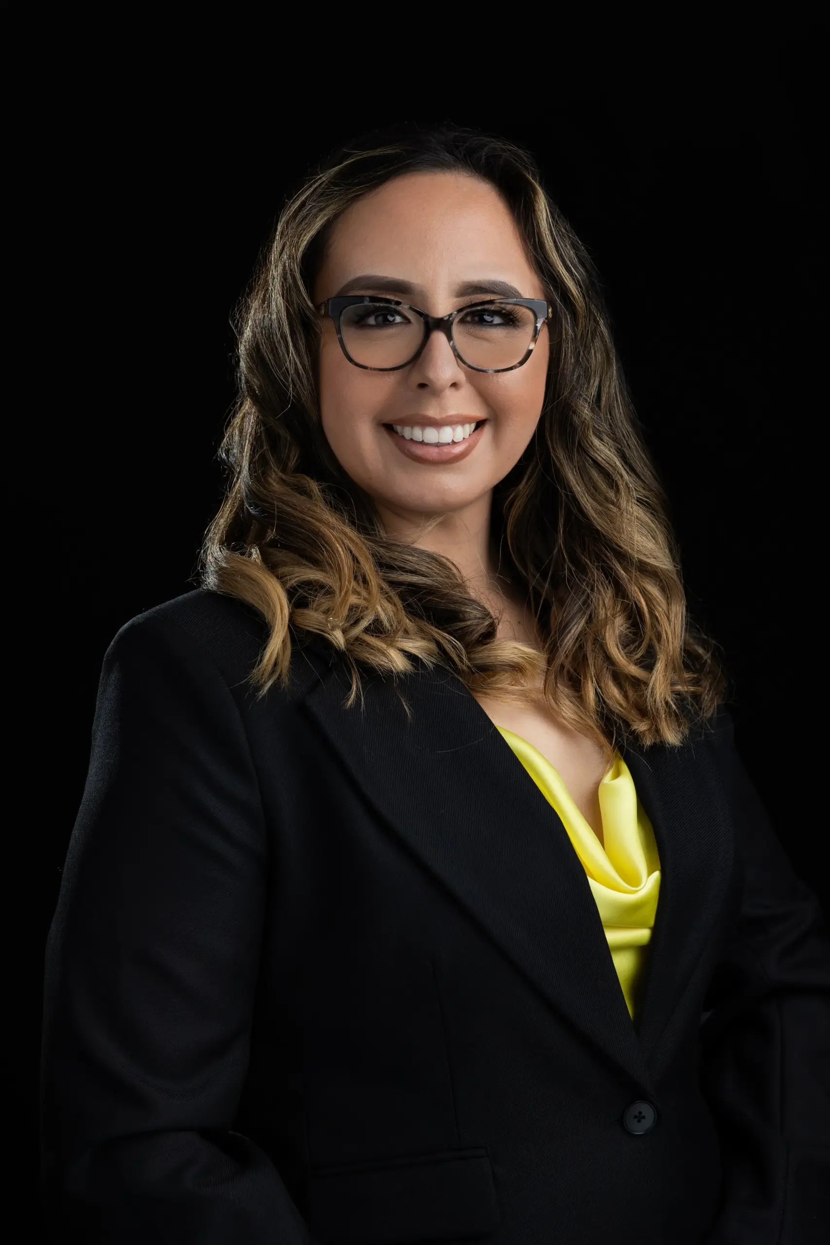Professional headshot of a woman in a black blazer and yellow top against a dark background